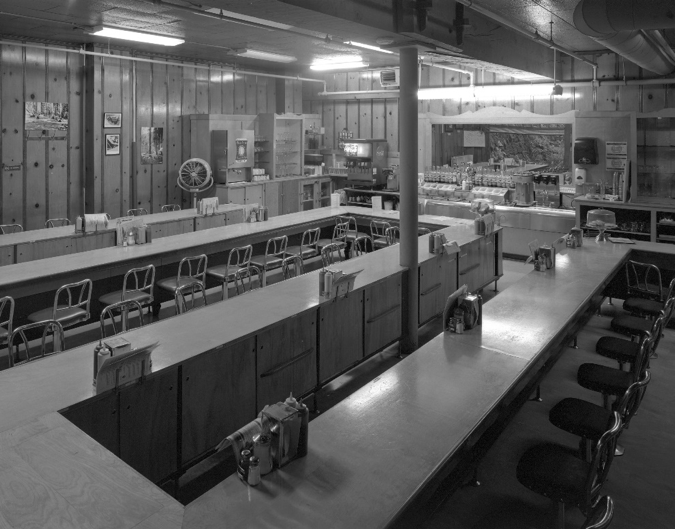 The coffee shop and lunch counter was completed about two years after construction. The post at center was added in 1954 to support the beam cracked when a flood swept through this room in 1936, shifting the whole building’s structure. Marks from debris from a second flood in 1964 are still visible on the original knotty pine paneling.